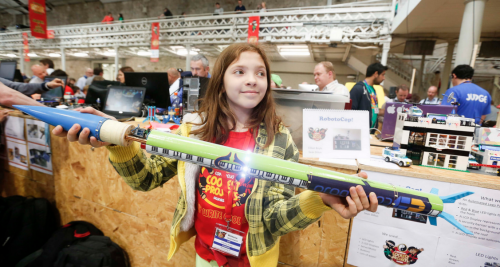 A girl in a CoderDojo Ninja T shirt proudly holds the rocket she has built; it's as long as she is tall