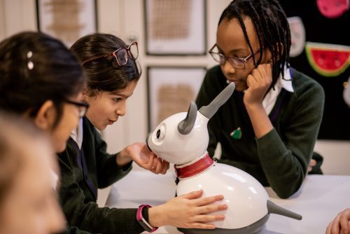 Three school children in uniforms stroke the robot dog's chin
