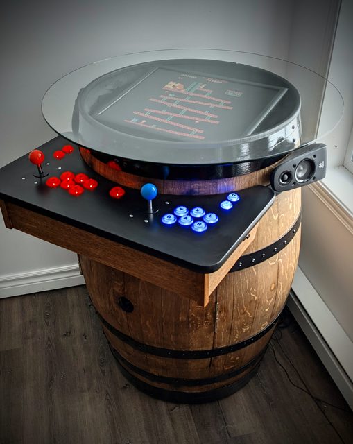 A beer barrel with drinks inside two opening doors cut into the front of the barrel and a retro arcade console serving as the lid of the barrel with joystick and buttons on a ledge in front 