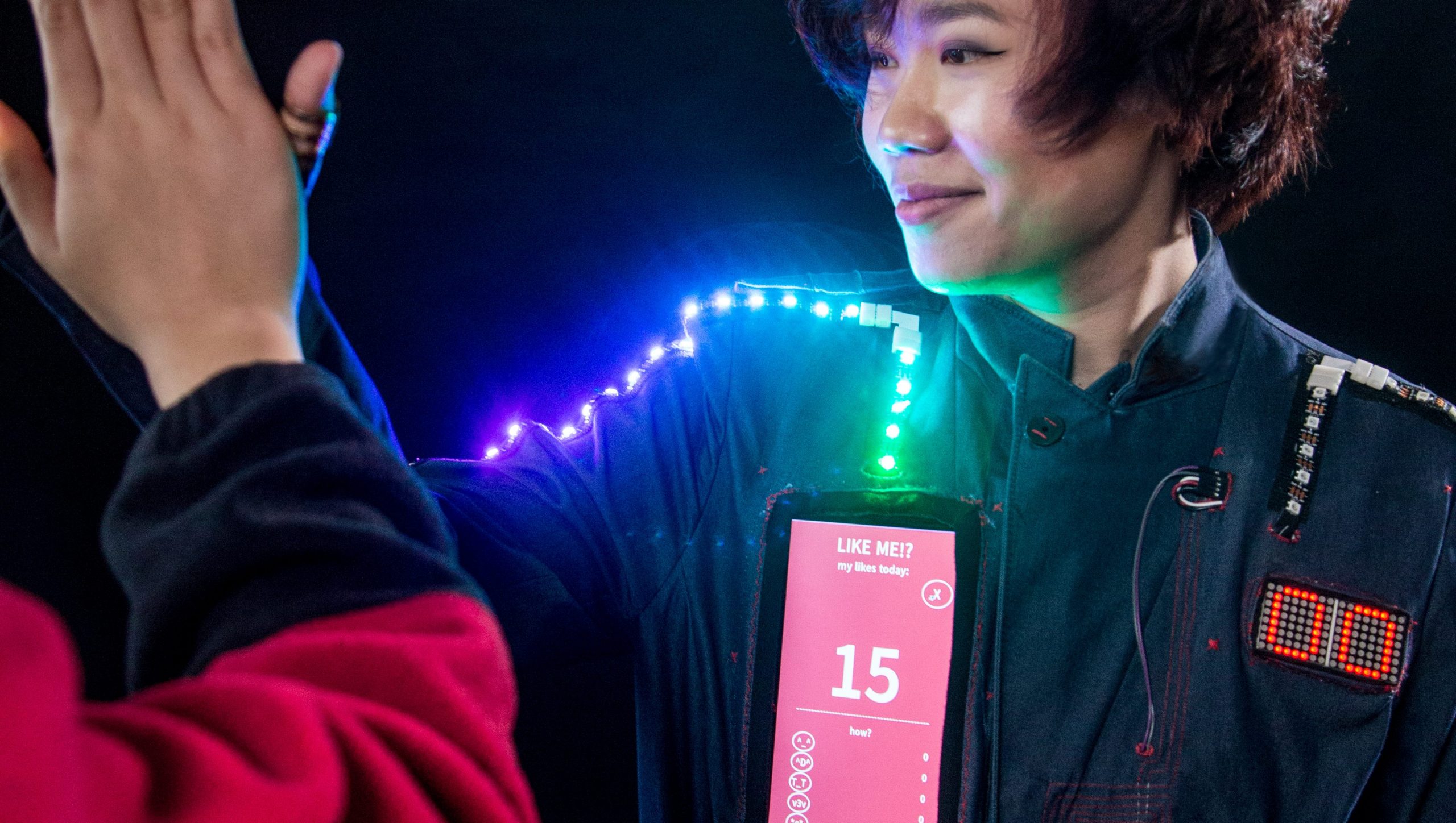 A MonthOfMaking project: Someone wearing a wearable tech project featuring LEDs, a two-digit LED matrix, and a tablet screen. The person is high-fiving someone who is out of view.