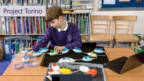 A boy creates a computer program using the Torino tool. There are several Torino pods attached to each other and the boy is using his hands to follow the sequence of the program as it runs.