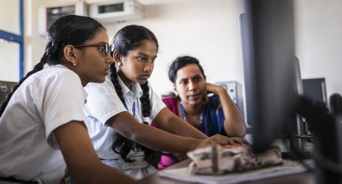 Two girls code at a desktop computer while a female mentor observes them.