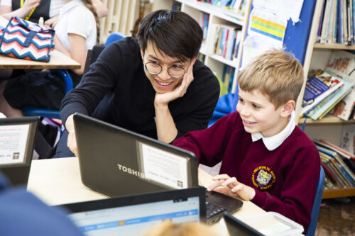 A primary school boy codes at a laptop with the help of an educator.