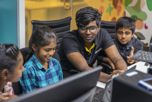 Three young people learn coding at laptops supported by a volunteer at a CoderDojo session.