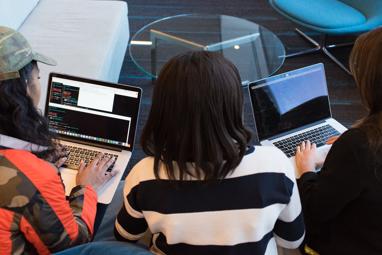 Three women sit on a sofa and work on laptops.