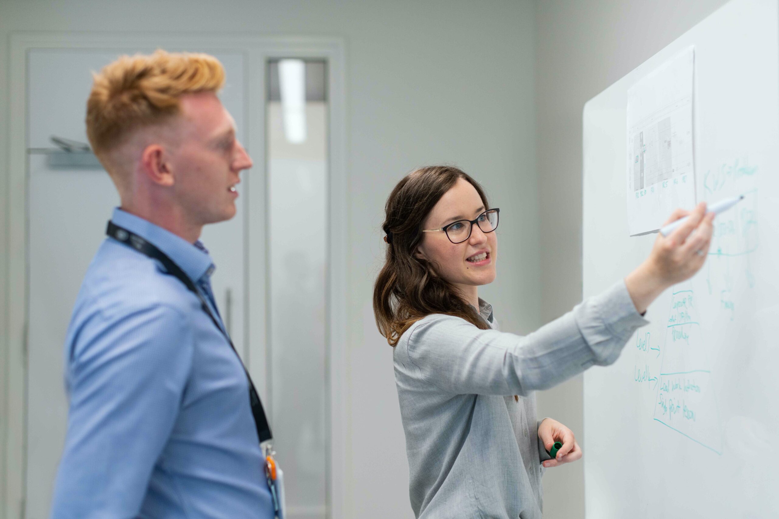 A woman explains something to a man at a whiteboard.