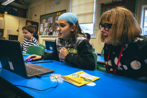 A girl codes on a laptop while a woman watches during a Code Club session.
