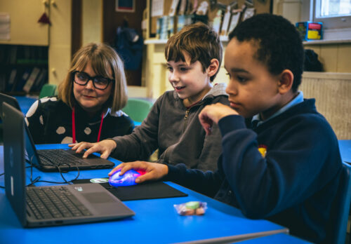 Two children code on laptops while an adult supports them.