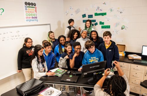 A group of teenagers pose for a group photo in a computer science classroom.